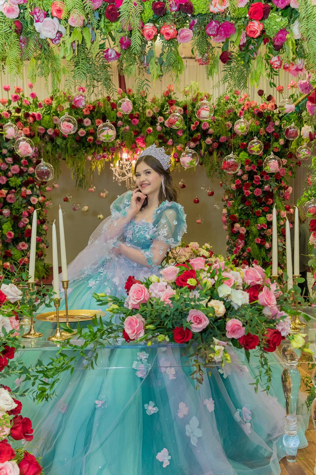 Quinceañera in turquoise gown seated under cascading floral arch at MP Venue